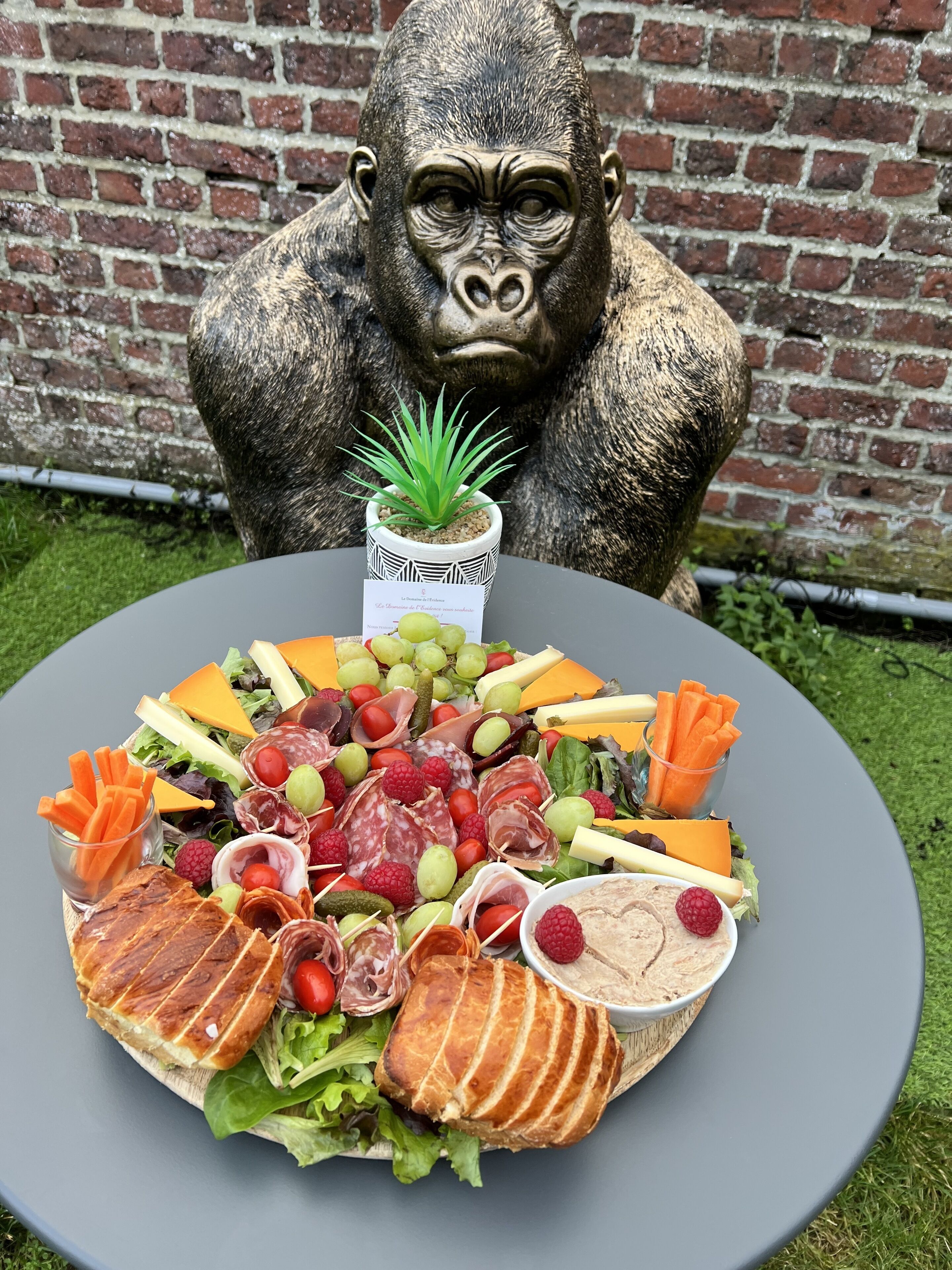 Une table ronde, dressée devant un mur de briques, présente une variété d’aliments, notamment des fromages, des viandes et des fruits. Une statue de gorille se trouve en arrière-plan.
