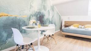 Ein modernes Zimmer mit einem runden Esstisch, zwei weißen Stühlen, einem Bett mit blauer Matratze und einem großen abstrakten Gemälde an der Wand.