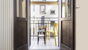 Ein offener Balkon mit gelbem Stuhl und kleinem Tisch, mit Blick auf ein Gebäude mit zahlreichen Fenstern und Balkonen.