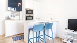 Eine moderne Küche mit weißen Schränken, einem eingebauten Ofen und einer Kaffeemaschine. Zwei blaue Barhocker und ein Fernseher auf einem weißen Schrank stehen dort.