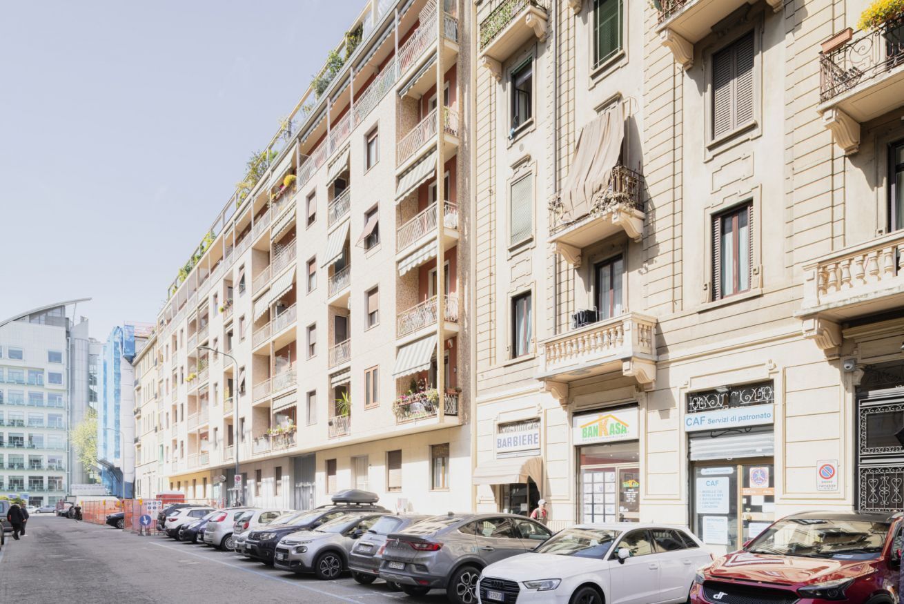 Une rangée de voitures garées le long d’une rue, avec un immeuble à plusieurs étages doté de balcons ornés de plantes, une devanture de magasin bien visible avec une enseigne, et un ciel dégagé.