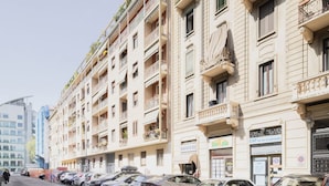 Eine Reihe geparkter Autos säumt eine Straße mit einem mehrstöckigen Gebäude, das Balkone mit Pflanzen, eine sichtbare Schaufensterfront mit einem Schild und einen klaren Himmel aufweist.