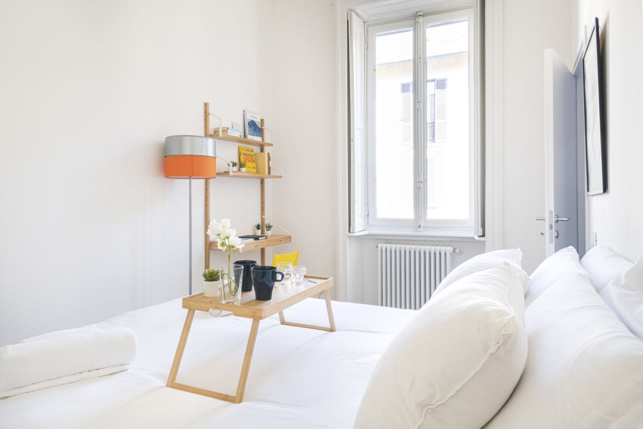 A bright, minimalist bedroom with a wooden bedside table, a lamp, and a window with a view of the outside.
