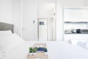 A modern hotel room with a neatly made bed, a small bedside table with a tray of plants, a microwave, and a kitchenette.