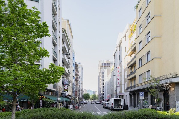A street view with multi-story buildings, parked vehicles, and a tree-lined sidewalk.