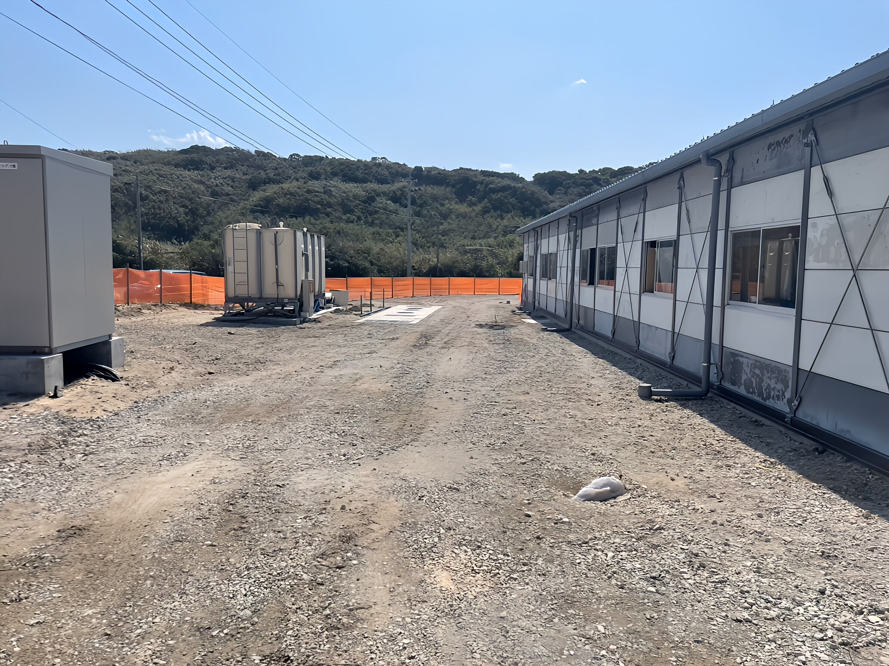 A construction site with a building under construction, a gravel ground, and a fenced area with trees in the background.