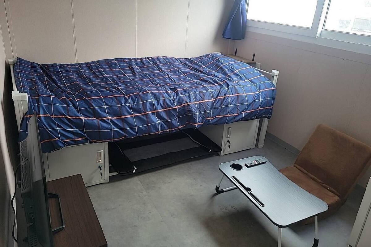 A single bed with a blue and white striped bedspread, a small table, a chair, and a window with a curtain.