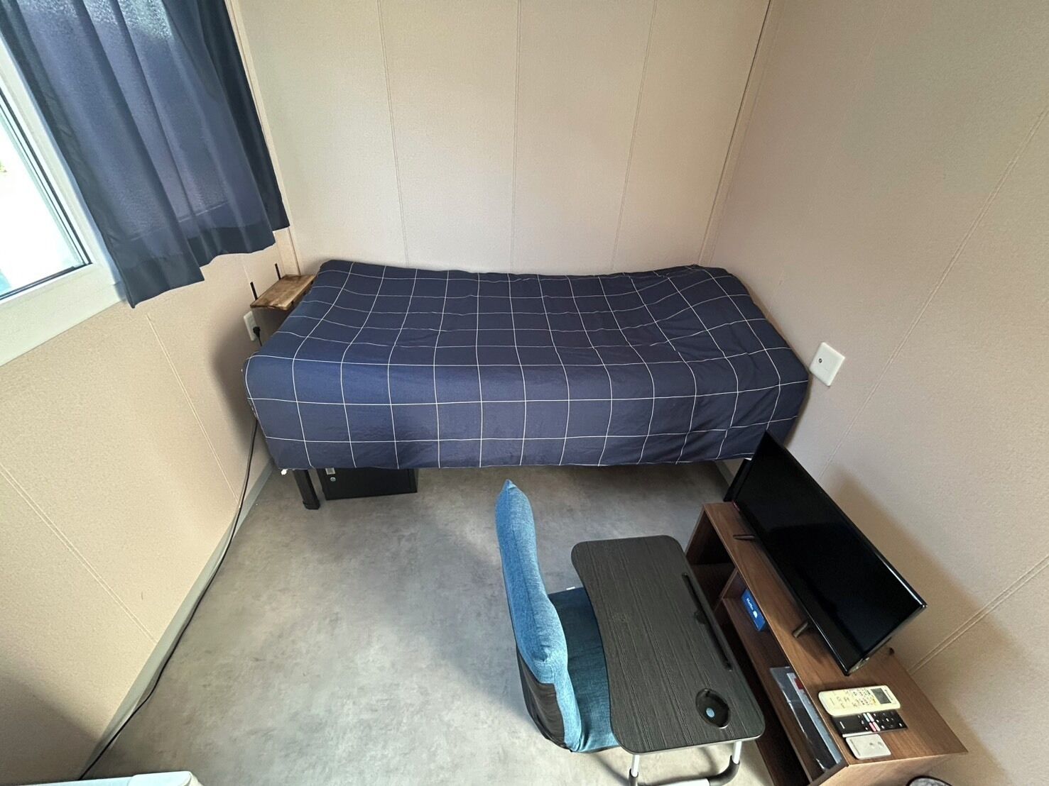 A single bed with a blue grid-patterned bedspread, a small bedside table with a television, and a window with curtains.