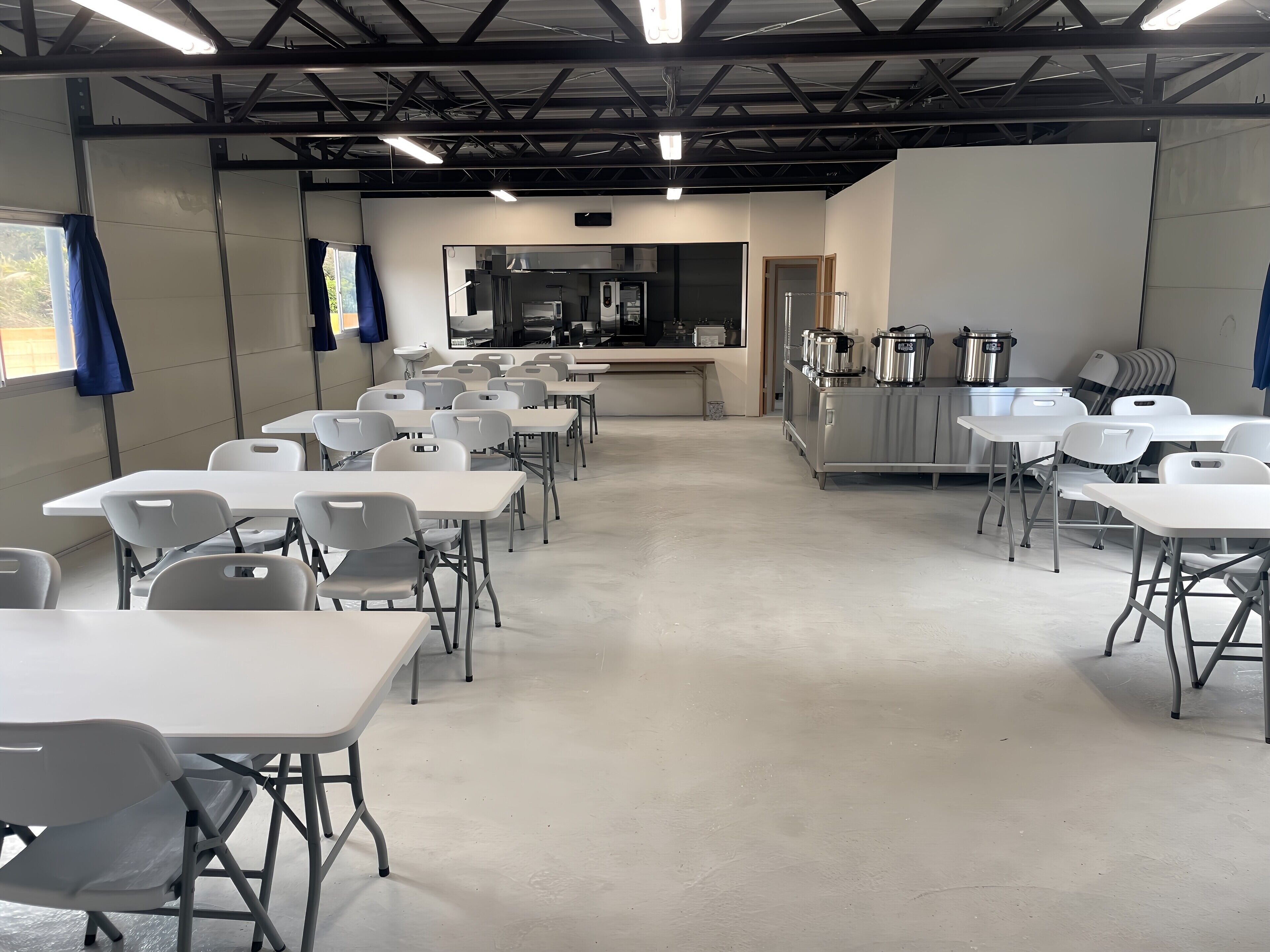 A classroom with white tables and chairs, a kitchenette area, and a large window with curtains.