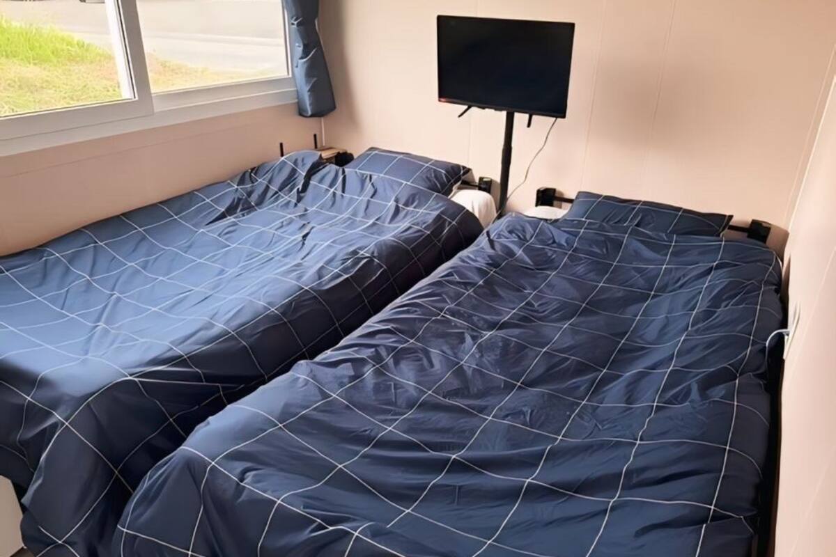 Two single beds with blue bedding, a flat-screen TV mounted on the wall, and a window with curtains.