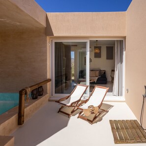 A sunlit balcony with two wooden lounge chairs and a small wooden table, a sliding glass door leading to an interior living area, and a small pool area. - Gaea Dream Villas Complex (Tinos)