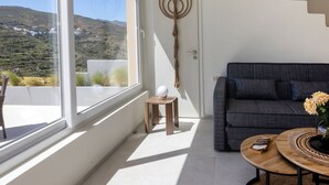 A modern living room with a grey sofa, a round wooden coffee table, and a view of a mountainous landscape through large windows. - Gaea Dream Villas Complex (Tinos)