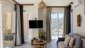 A modern living room with a large TV, a round wooden table, a sofa, and a view of the countryside through large windows. - Gaea Dream Villas Complex (Tinos)