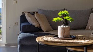 A modern living room with a grey sofa, a round wooden coffee table, and a bonsai tree on the table. - The Hydor Element Of Gaea (Tinos)