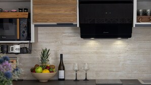 A modern kitchen with wooden cabinets, a microwave, a built-in oven, a fruit bowl, wine glasses, and a bottle of wine. - The Hydor Element Of Gaea (Tinos)