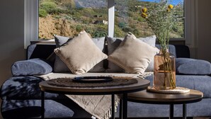 A modern living room with a sofa, pillows, a round table, and a vase with flowers. A large window offers a view of a mountainous landscape. - The Pyr Element Of Gaea (Tinos)
