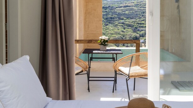 A balcony with a table and chairs, a view of a hilly landscape, and a bed with white bedding. - The Pyr Element Of Gaea (Tinos)
