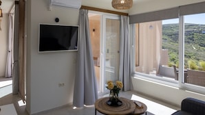 Una sala de estar moderna con un televisor de pantalla plana, una mesa de centro redonda con un jarrón de flores, un sofá gris y un ventanal con vistas a un paisaje montañoso.