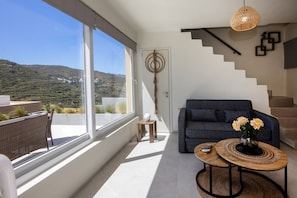 A modern living room with a large window, a sofa, a coffee table, and a staircase. - The Aether Element Of Gaea (Tinos)