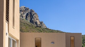 Un edificio con fachada color beige, una ventana de marco blanco y una puerta. Se observa una colina rocosa al fondo bajo un cielo azul despejado.