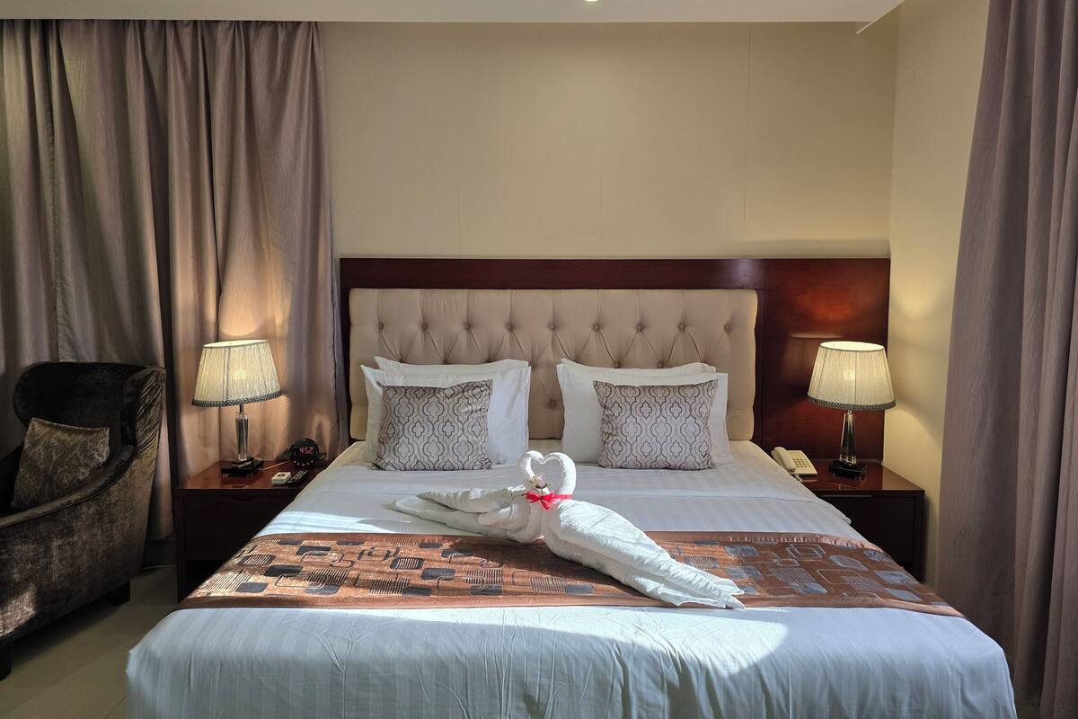 A neatly made bed with a decorative towel, two patterned pillows, and a headboard. There are two bedside tables with lamps and a chair to the left.