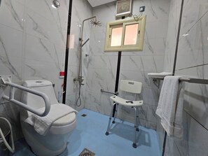 A bathroom with a toilet, a shower, a handrail, a wheelchair-accessible chair, and a towel rack. - Dar Al Wedad Serviced Apartments (Jeddah)