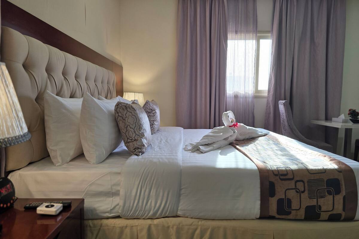 A hotel room with a large bed, a wooden headboard, a bedside table with a lamp, a clock, and a small table with a plant.