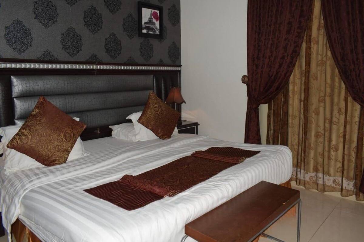 Habitación de hotel con una cama grande, dos almohadas, una mesita de noche con lámpara y un cuadro en la pared.