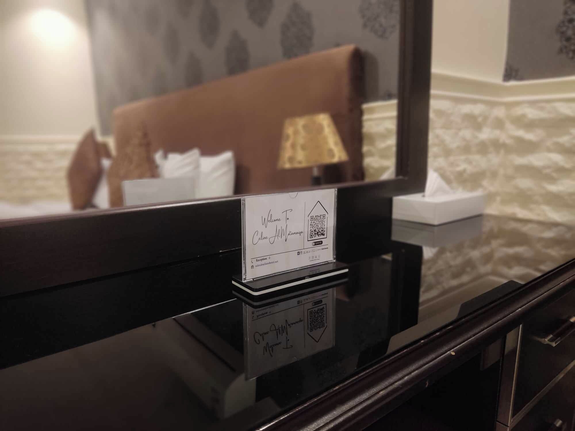 Un mostrador de recepción de hotel con una tarjeta de recepción, un espejo que refleja una cama y una lámpara, y una pared con textura al fondo.