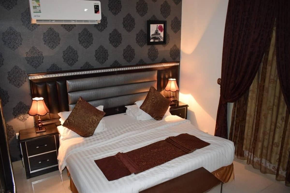 Una cama bien hecha con una colcha blanca y almohadillas decorativas marrones, flanqueada por dos mesitas de noche con lámparas, en una habitación con papel tapiz estampado y un cuadro enmarcado en la pared.