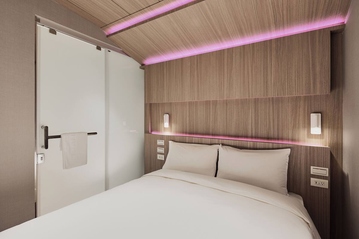 Moderni hotellihuone, jossa on puinen pääty, valkoinen sänky ja ovi, jonka kahvassa roikkuu pyyhe.