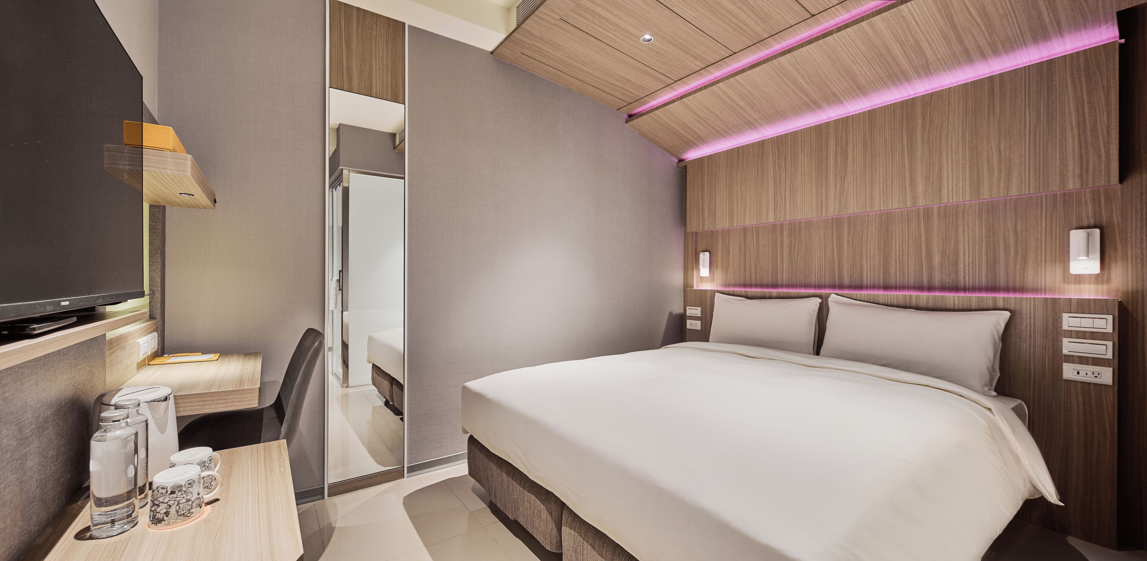 A modern hotel room with a large bed, a desk with a chair, a television, and a mirror.