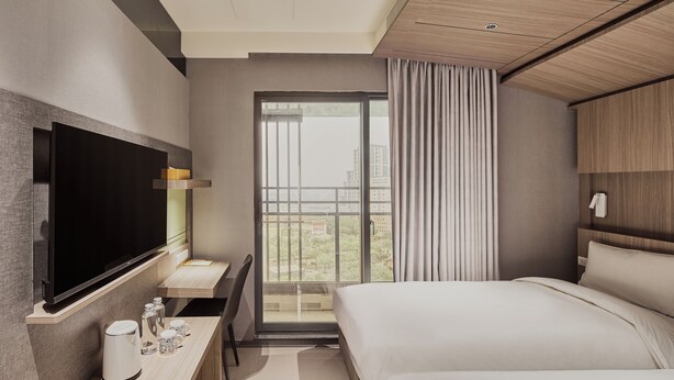 Een moderne hotelkamer met een groot bed, een flatscreen televisie aan de muur, een bureau met een koffiezetapparaat en uitzicht op de stad door een groot raam.