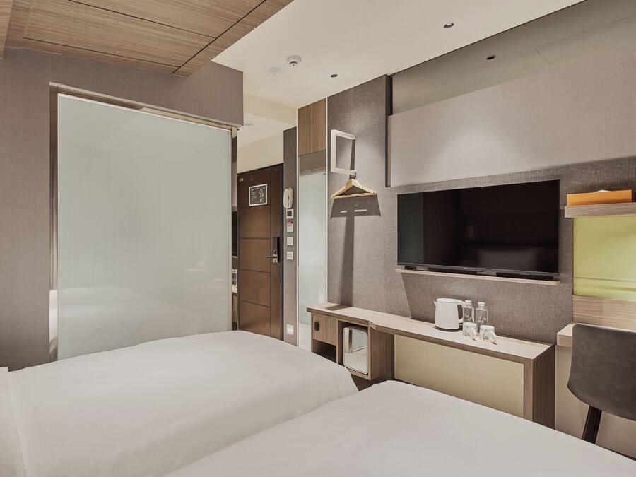 A hotel room with two beds, a desk with a TV, a mirror, and a small kitchenette area.