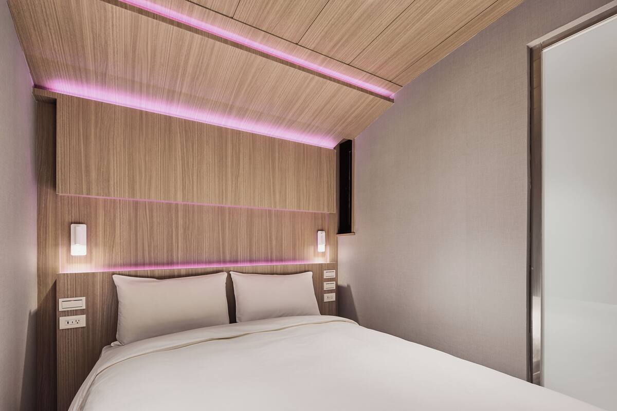 Moderni hotellihuone, jossa on puinen sängynpääty, yöpöytävalaisimet ja neutraali värimaailma.