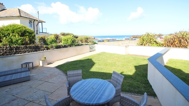 A patio with a round table and wicker chairs, overlooking a grassy area and the sea. - Spacious dog friendly apartment by the beach, private garden and sea views. (Padstow)