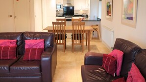A living room with a brown leather sofa and a dining area with wooden chairs and a table. - Spacious dog friendly apartment by the beach, private garden and sea views. (Padstow)