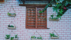 A brick wall with a window featuring a wooden shutter. There are five hanging planters with green plants. - Nama Stay Home (Pokhara)