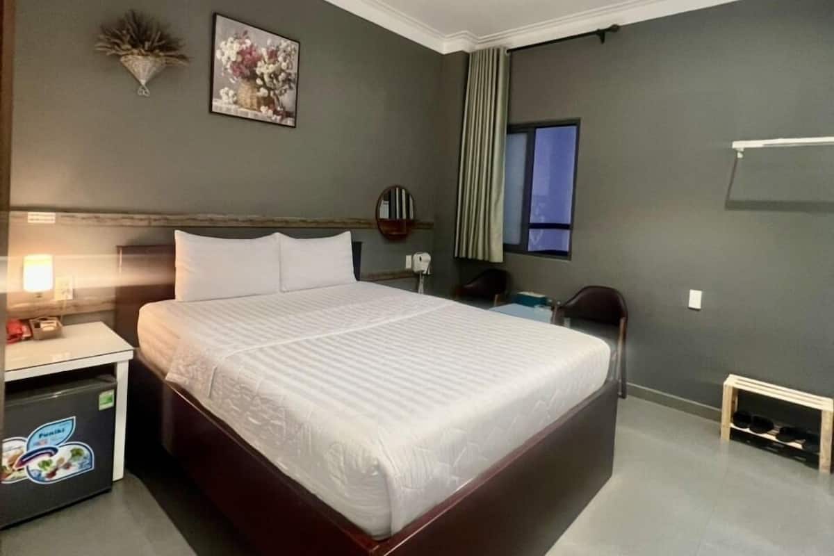 Ein modernes Hotelzimmer mit einem großen Bett, einem Nachttisch, einem Sessel, einem kleinen Tisch, einem am Wand montierten Fernseher und einem Fenster mit Vorhängen.