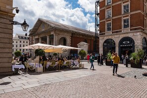 Ein Straßencafé mit Tischen und Sonnenschirmen, ein historisches Gebäude mit Bögen und ein modernes Gebäude mit einem Schild.