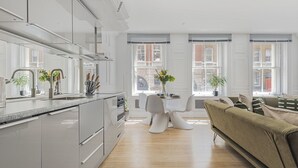 Una cocina moderna con armarios blancos, un comedor con mesa y sillas, y un sofá en un salón luminoso con ventanas amplias.