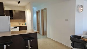 A modern kitchen with dark wooden cabinets, a white countertop, and a built-in dishwasher. There is a bar stool near the counter and a dining table with chairs in the foreground. - Spacious 2-bed Apartment in Sotira With Pool (Sotira)