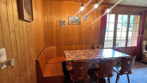Un interior de madera con una mesa de comedor, sillas y un banco. Hay cuadros en la pared y una ventana con cortinas.