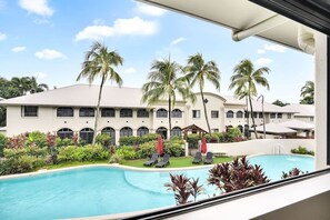 Vista de la piscina con un edificio de fachada blanca y ventanas arqueadas, rodeado de palmeras y vegetación exuberante.