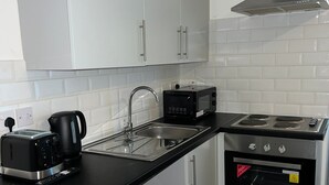 A compact kitchen with white cabinets, a black countertop, a built-in microwave, a kettle, a toaster, and a stove. - Comfort Studio Ensuite (Skegness)