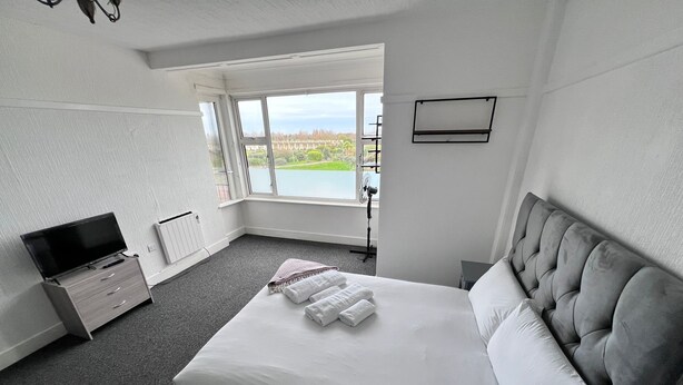 A hotel room with a large bed, a television on a dresser, a window with a view of a park, and a fan near the window. - Comfort Studio Ensuite (Skegness)