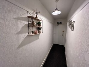 A hallway with a white textured wall, a black floor, and a white door with a fire exit sign. There are shelves with plants and decorative items, and a pendant light hanging from the ceiling. - Two Bedroom - Apartment (Skegness)