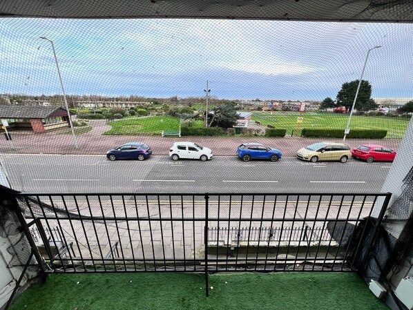 A view from a balcony with a metal railing, overlooking a parking lot with several cars and a green sign in the background. - Three Bedroom - Apartment ( Sea View ) (Skegness)