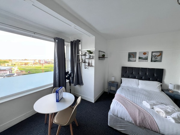 Hotellihuone, jossa on sänky, pieni pöytä ja tuoli, yöpöytä lampun kanssa, seinälle kiinnitetty hylly koriste-esineitä varten sekä ikkuna, jossa on verhot.