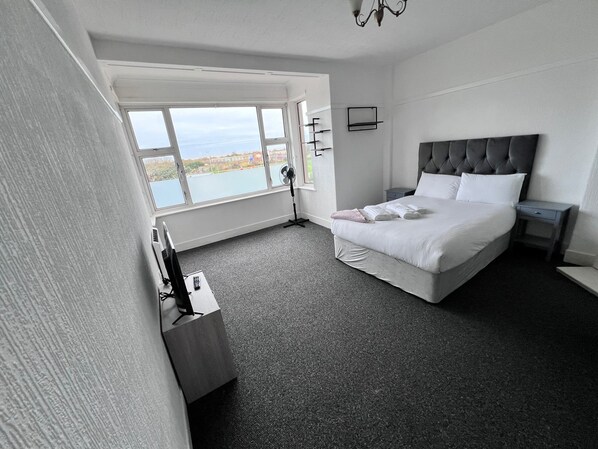A hotel room with a large window, a bed with white linens, a bedside table, and a wall-mounted fan. - Three Bedroom - Apartment ( Sea View ) (Skegness)